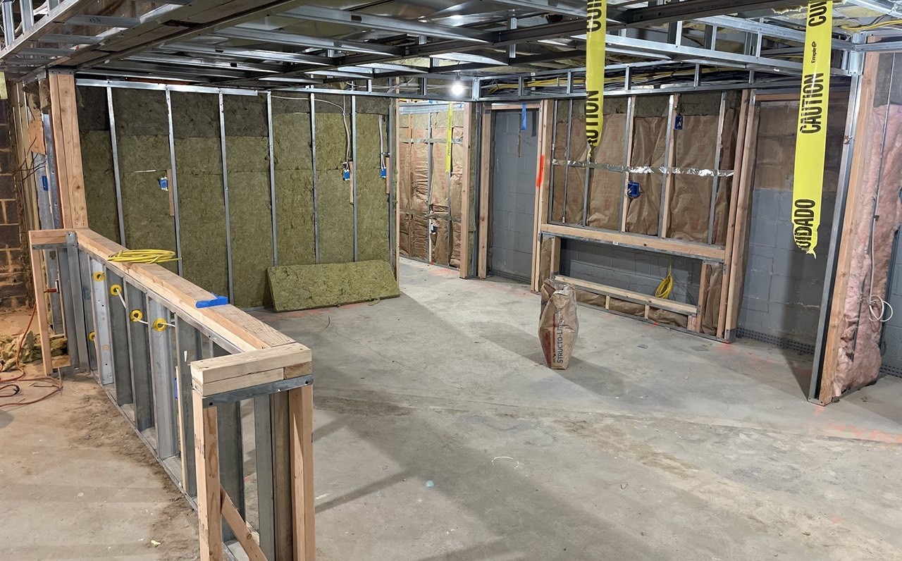 An unfinished basement under construction showing exposed wall studs, yellow caution tape, insulation, a bare concrete floor, and the rough timber framing for the future bar and fireplace layout.