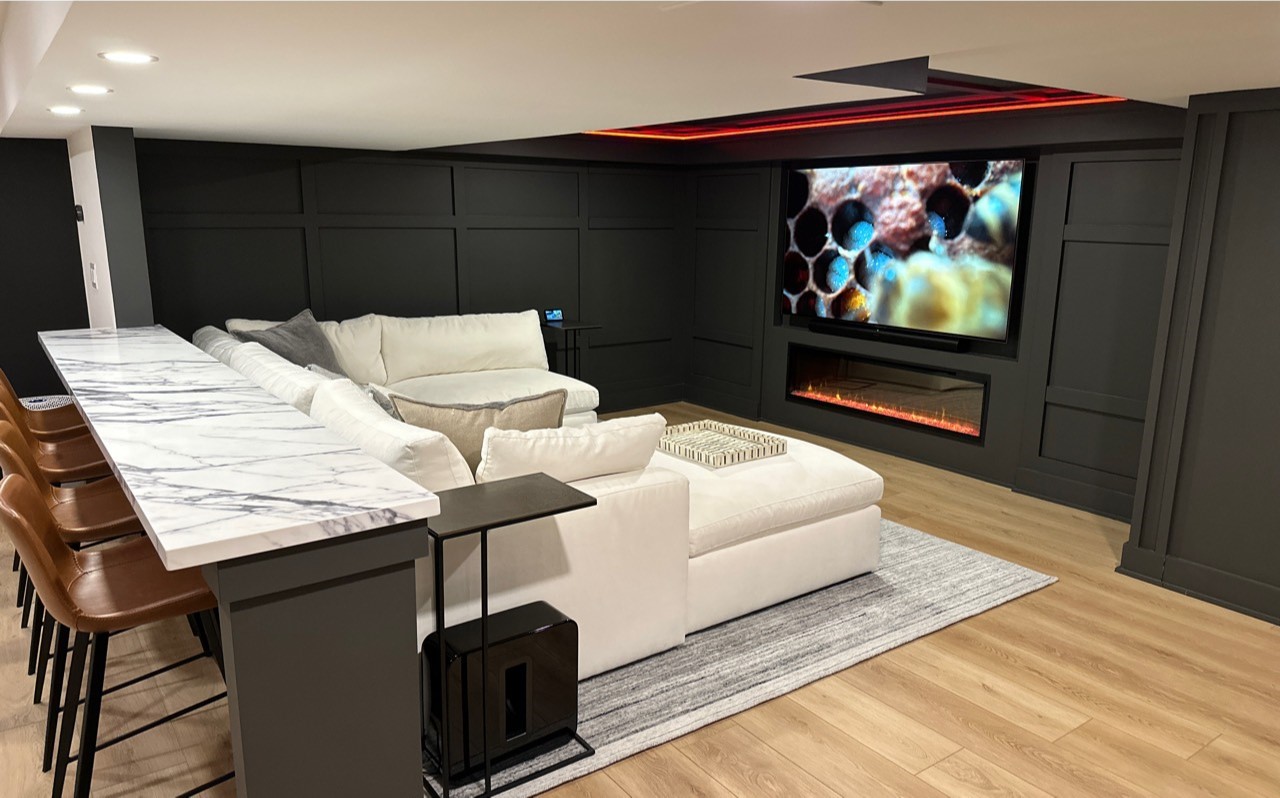 A modern, finished basement home theater and bar featuring dark paneled walls, a plush white sectional sofa facing a mounted TV and electric fireplace, and a marble-topped bar with leather stools.